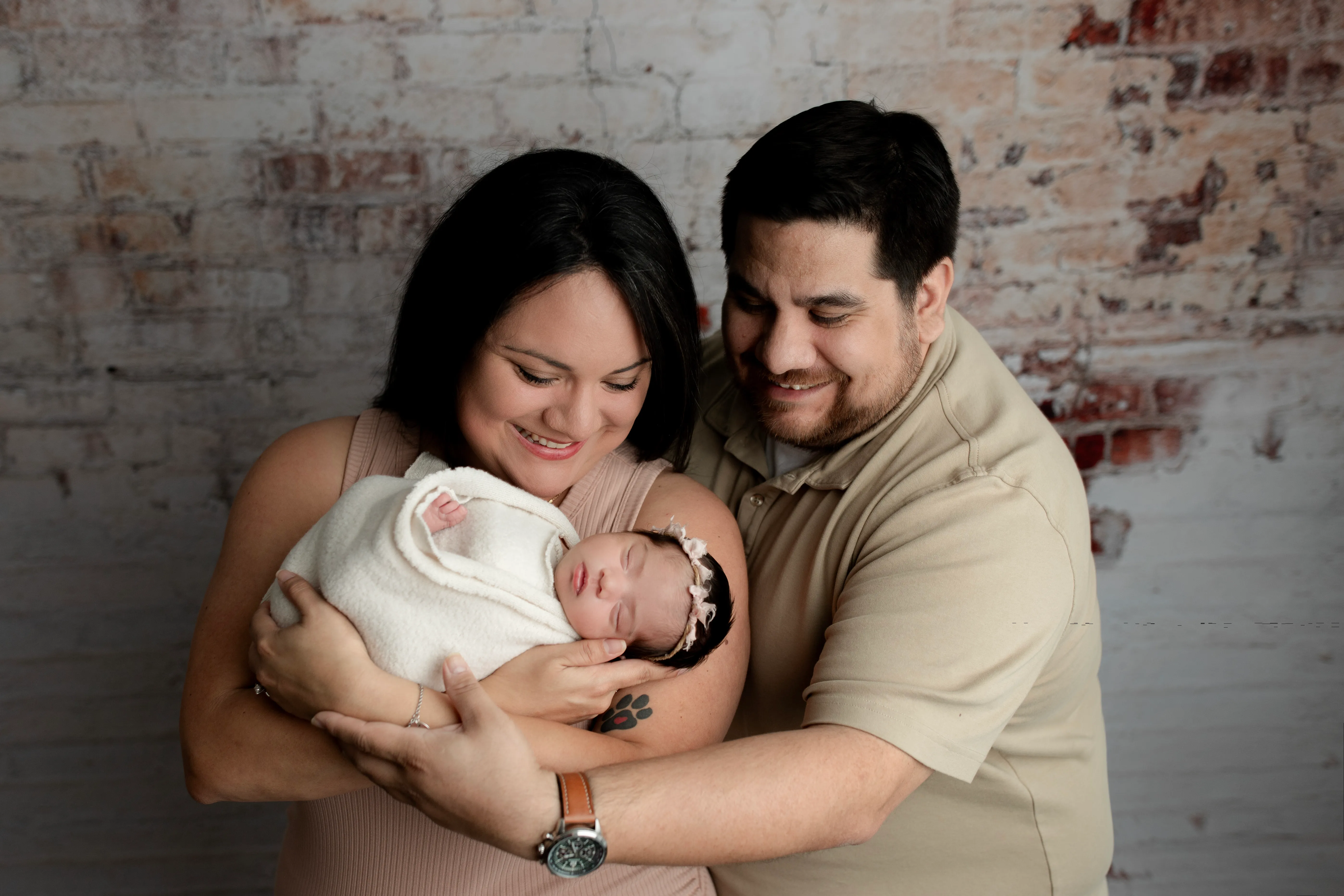 Alex and his wife holding their swaddled newborn daughter against a whitewashed brick wall, both looking down at her.